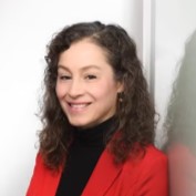 Person mit lockigem Haar in rotem Blazer und schwarzem Rollkragenpullover lächelt vor hellem Hintergrund.