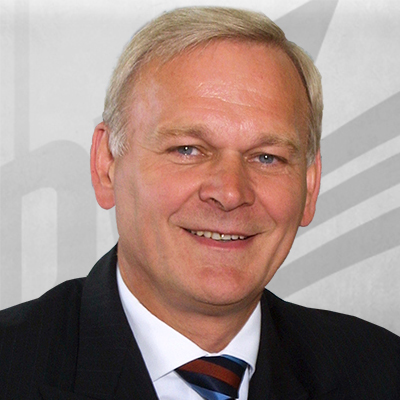 Ein lächelnder Mann mit hellem Haar in Anzug und Krawatte vor einem abstrakten Hintergrund.