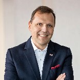 Ein lächelnder Mann in Anzug mit verschränkten Armen vor weißem Hintergrund.