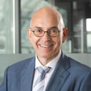 Eine lächelnde Person mit Brille, Glatze und Anzug vor einem unscharfen Hintergrund.