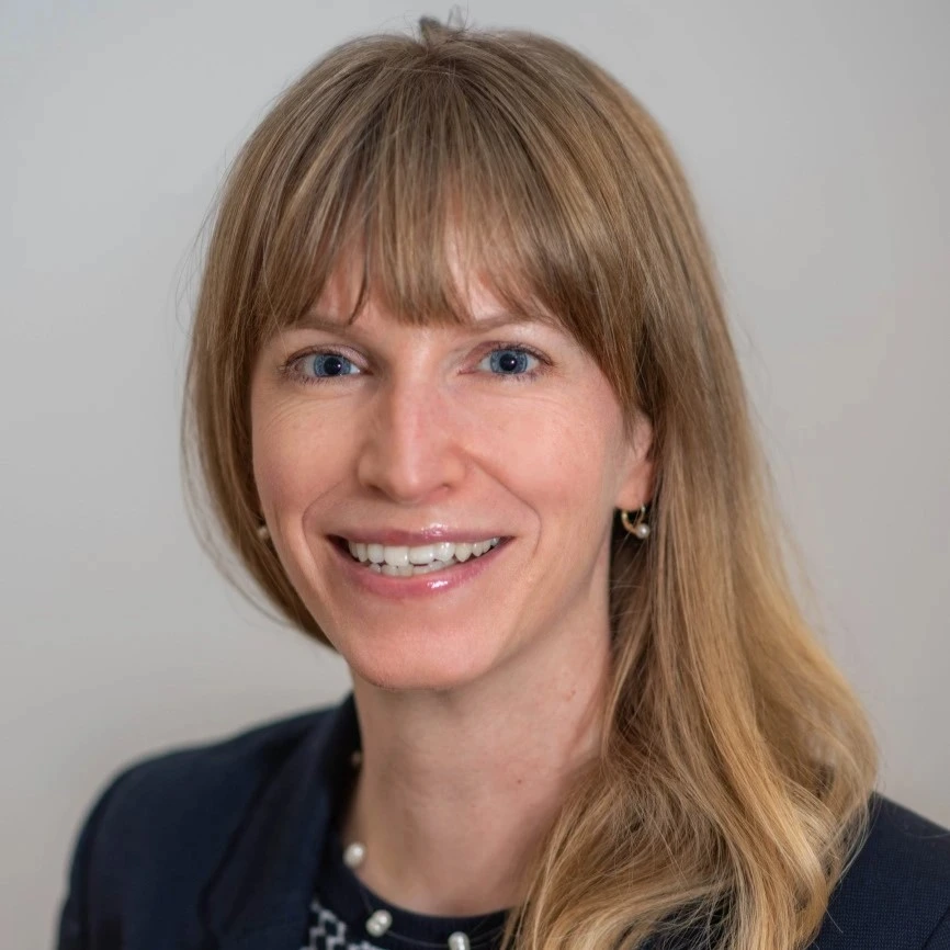 Porträt einer lächelnden Frau mit blonden Haaren und blauem Blazer vor neutralem Hintergrund.