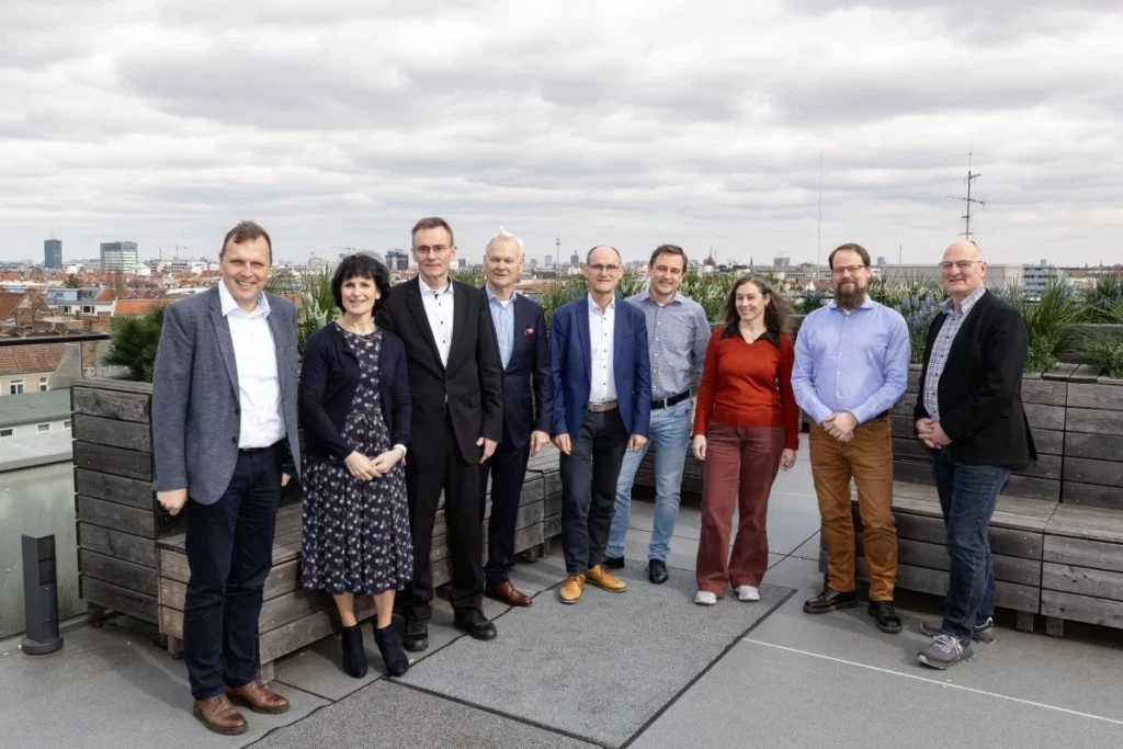 Neun Personen stehen auf einer Dachterrasse in urbaner Umgebung bei bewölktem Himmel.