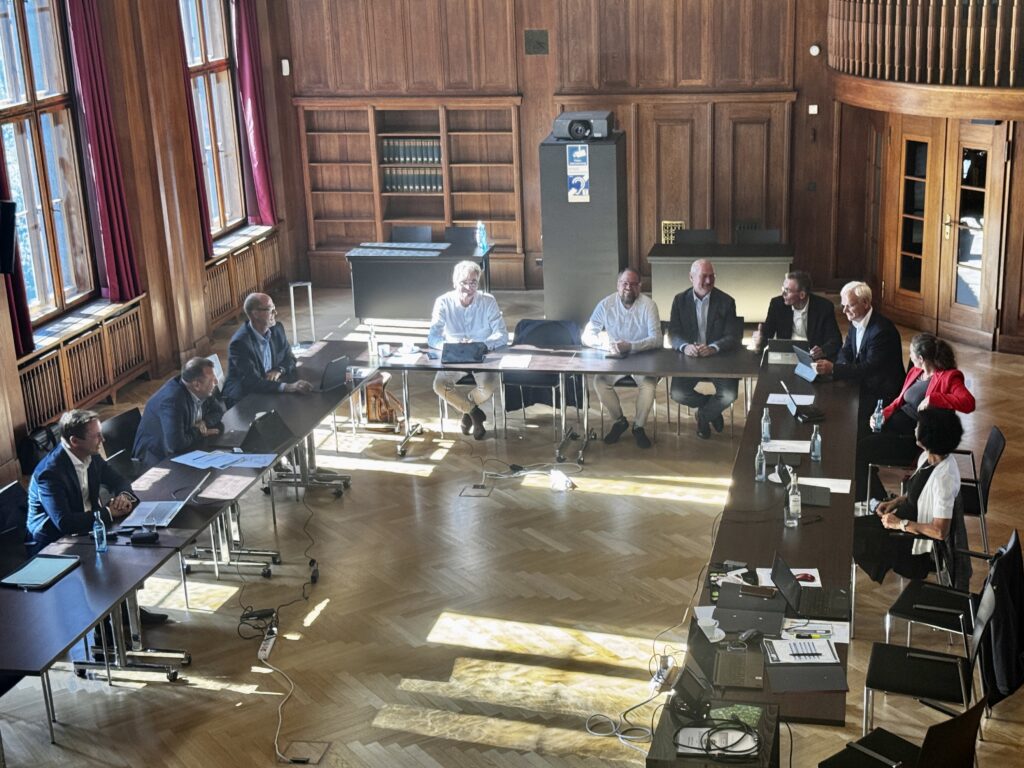 Menschen sitzen in einem großen, holzgetäfelten Raum an Tischen in U-Form während einer Besprechung.