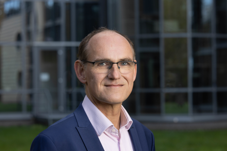 Person in Anzug und Brille steht vor einem gläsernen Bürogebäude.