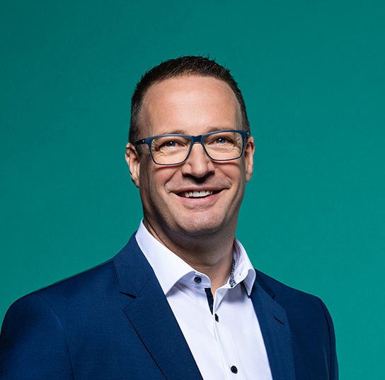 Mann mit Brille und Anzug vor einem türkisfarbenen Hintergrund, lächelt.