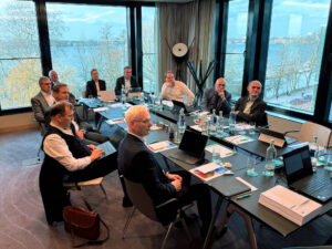 Mehrere Personen in einem Konferenzraum an einem rechteckigen Tisch, mit Laptops und Papieren, Blick auf einen See.