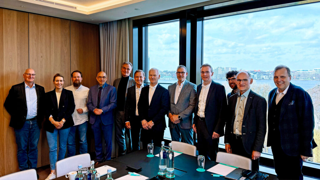 Gruppe von 13 Personen in Businesskleidung steht in einem Konferenzraum vor einem Fenster mit Stadtblick.