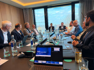 Eine Gruppe von Personen in formeller Kleidung bei einem Meeting an einem langen Konferenztisch mit Laptops und Getränken.